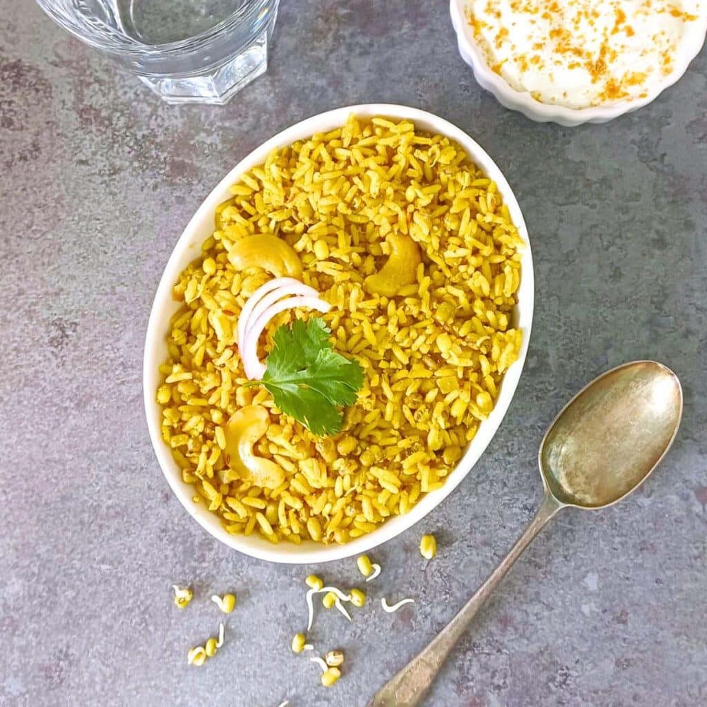 white bowl of moong sprouts pulao placed on a tile with a spoon and bowl of raita.