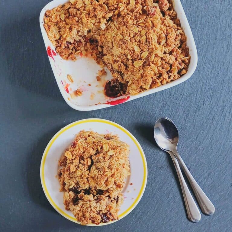 a plate of cherry crumble placed on black stone along with spoons and a baking dish.