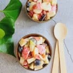2 bowls of fruit salad with cream placed on a tile with wooden spoons.