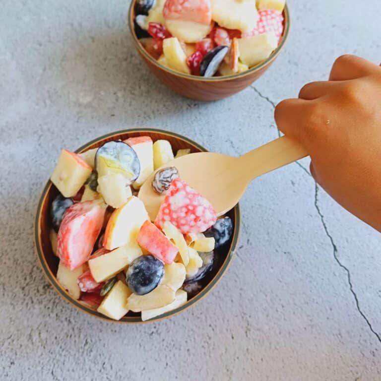 child scooping a spoon of fruit salad from the bowl placed on tile.