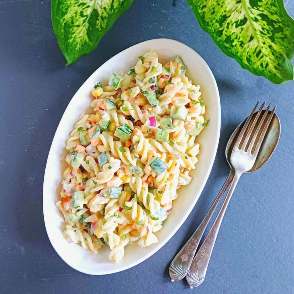 white oval bowl of vegetable pasta salad placed on blue cloth with a spoon and fork.