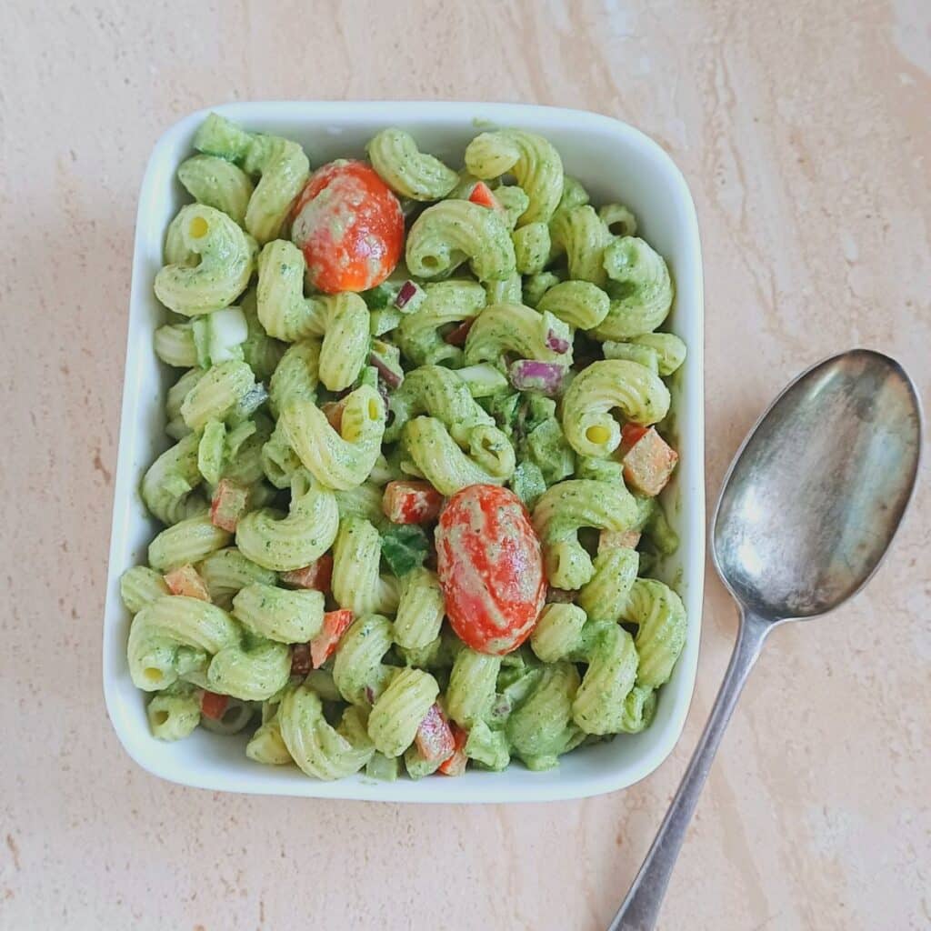 basil vegetable pasta salad in a square bowl placed on a tile along with a spoon.