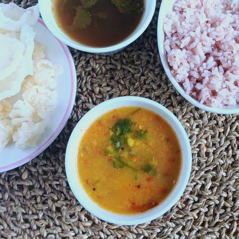 white bowl of toor dal placed on placemat along with a bowl of rice, rasam and papad.