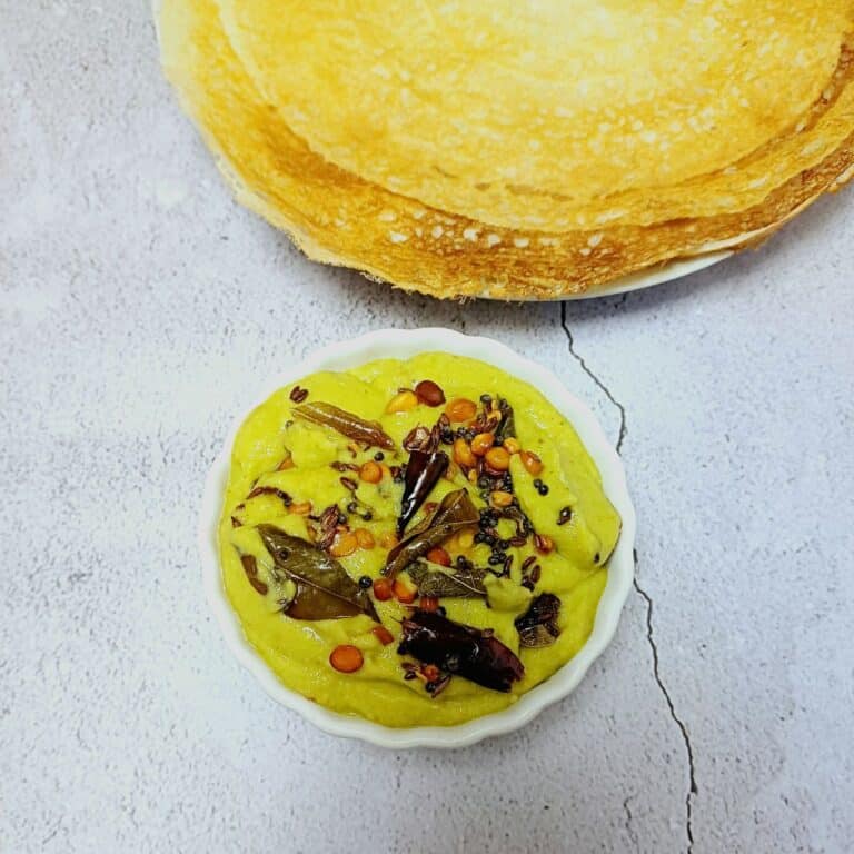 bowl of green tomato chutney placed on blue tile along with a plate of dosas.
