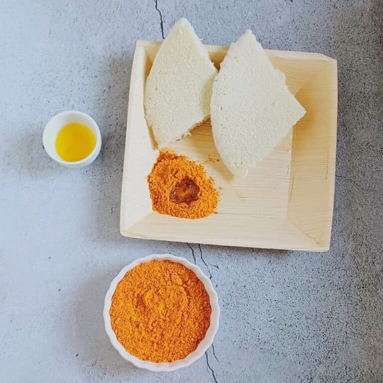bowl of idli podi placed on blue backdrop along with a pinch bowl of ghee and a square areca plate filled with 2 idlis, podi and ghee.