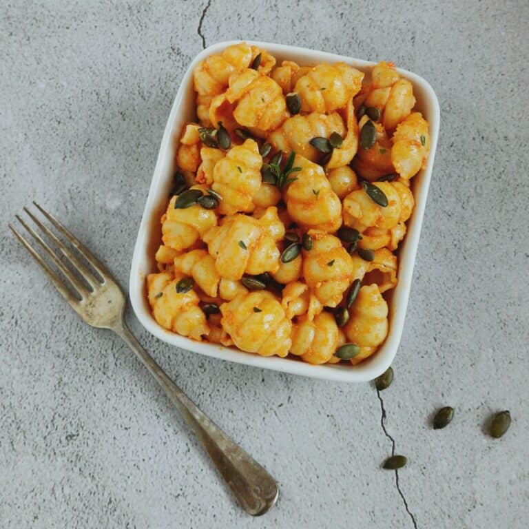 square bowl of roasted pumpkin pasta topped with pumpkin seeds placed on a tile along with a fork.