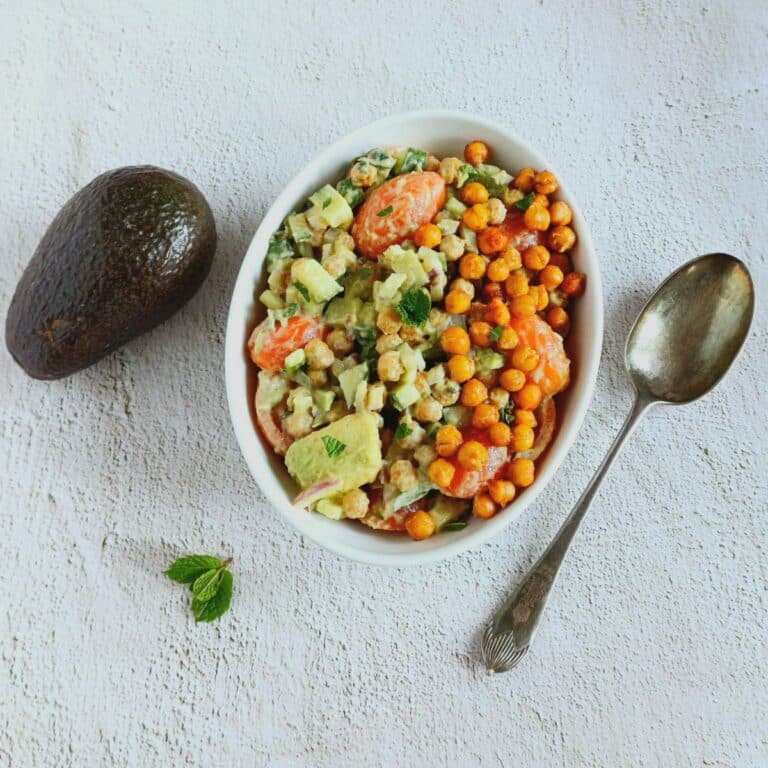 bowl of roasted chickpea vegetable salad placed on a tile along with a spoon and avocado.