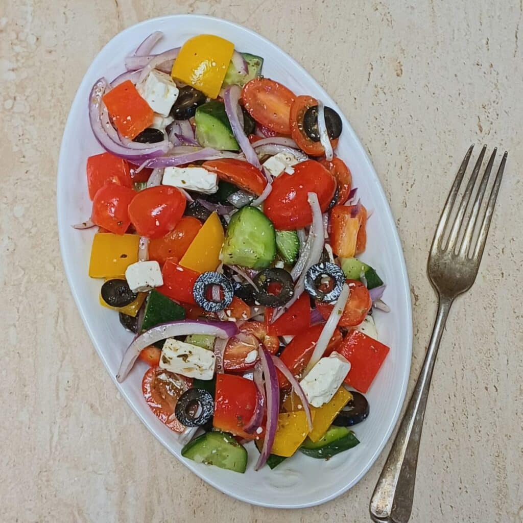 white plate of greek vegetable salad placed on granite along with a fork.