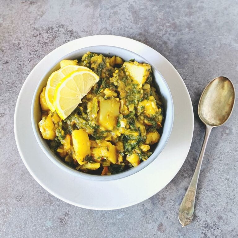 grey bowl of spinach potato curry with lemon slices on grey plate.