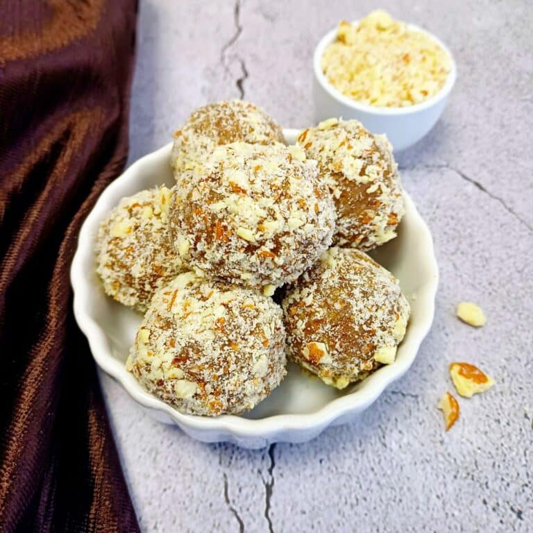 white bowl of fig energy balls placed on tile along with a pinch bowl of chopped almonds and brown cloth.