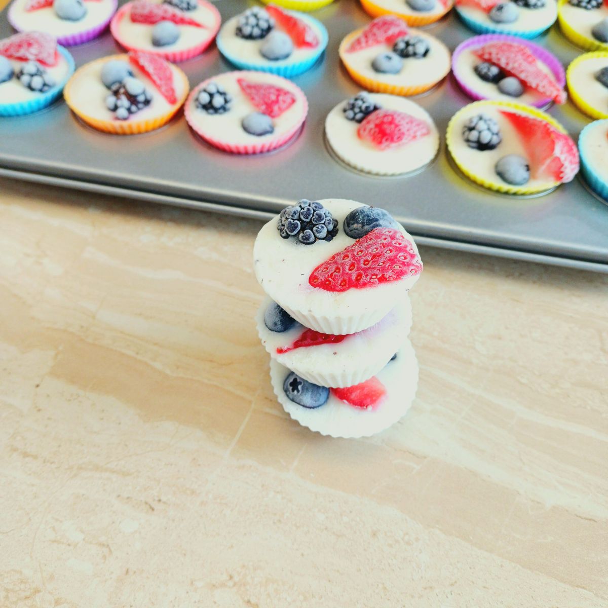 stacked frozen berry yogurt bites on a brown tile with more of them in a muffin tin behind.