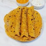 rolled up masala rotis placed on top of stack of rotis on a plate along with a bowl of raita and a bowl of curry on marble.
