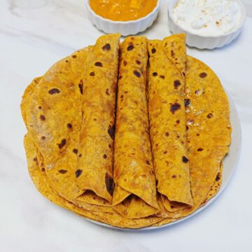 rolled up masala rotis placed on top of stack of rotis on a plate along with a bowl of raita and a bowl of curry on marble.