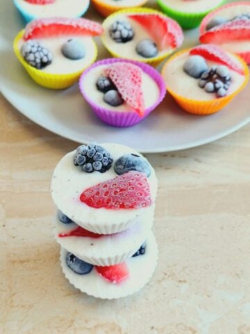 three stacked frozen mixed berry yogurt bites placed on a tile along with a grey plate of more frozen bites.