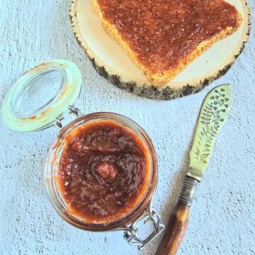 glass jar of 4 ingredient strawberry rhubarb jam placed on a tile along with a knife and toast spread with jam.