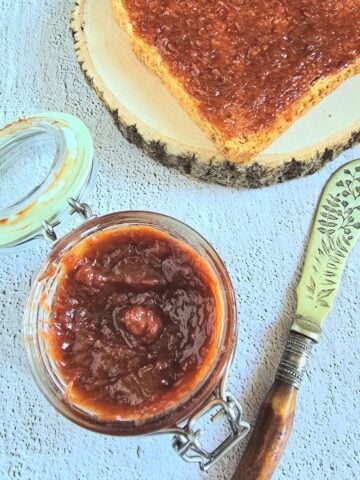 glass jar of 4 ingredient strawberry rhubarb jam placed on a tile along with a knife and toast spread with jam.