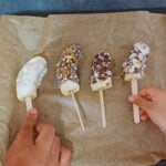 two children holding frozen banana pops placed on lined baking tray.