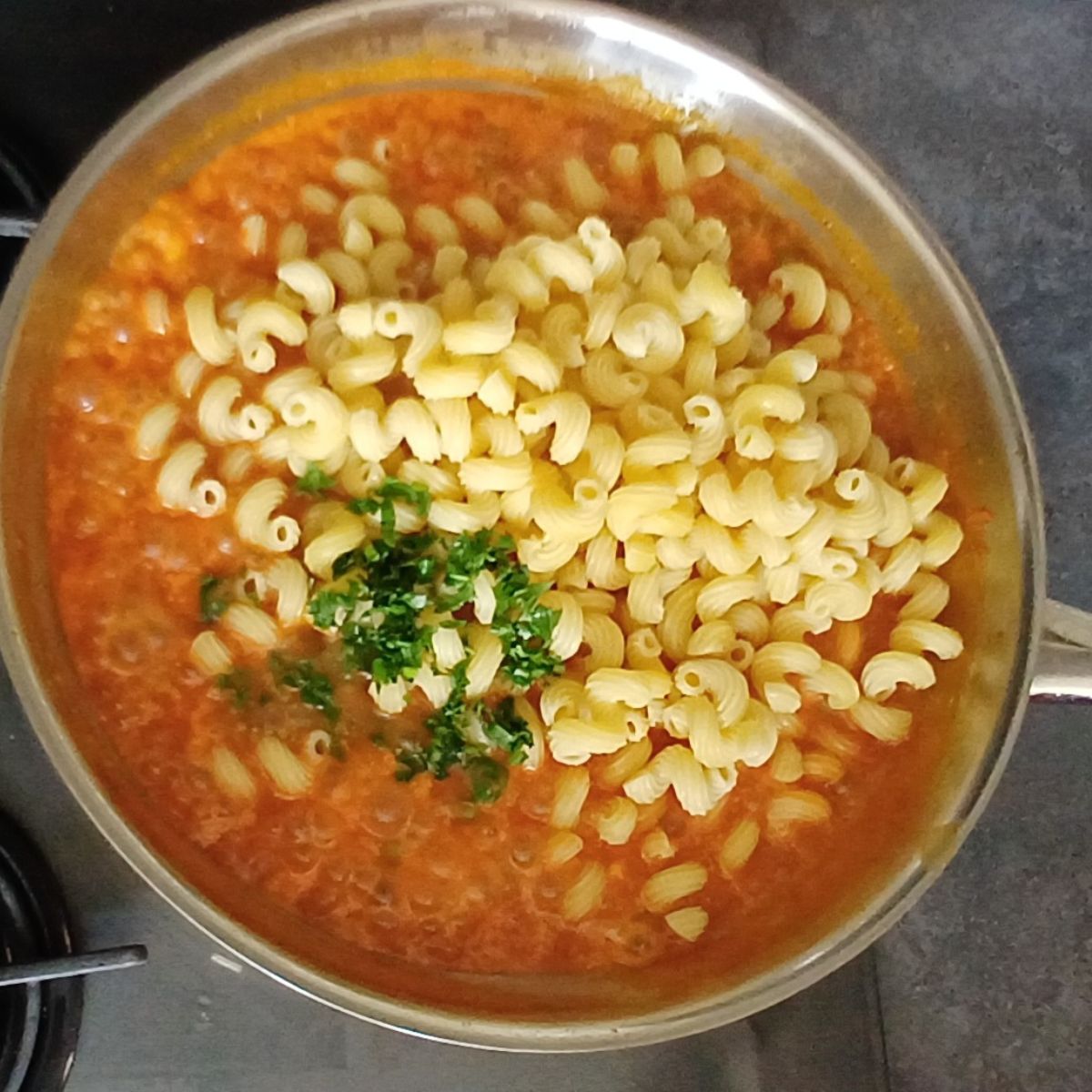 cooked pasta and green herbs on red vegetable sauce in a steel pan.