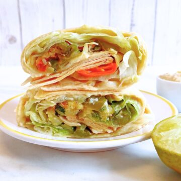 roast vegetables wrap with hummus on a white plate placed on marble along with half lemon and a bowl of hummus.