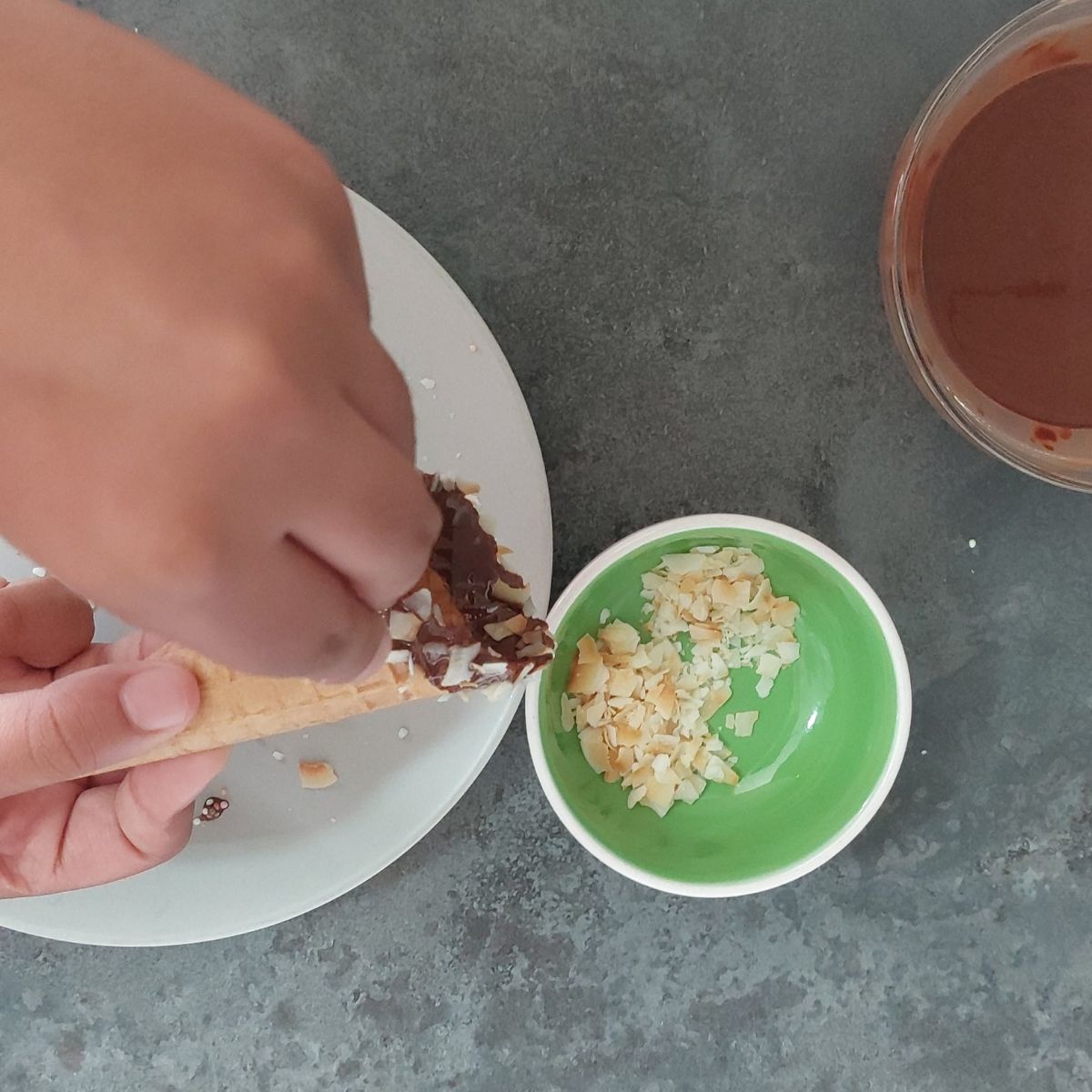 a child sprinkling toasted coconut flakes on chocolate coated ice cream cone. 