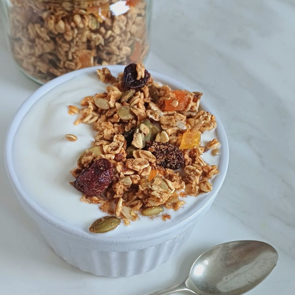 white bowl of yogurt topped with oil free granola placed on marble with a spoon next to it and a jar of granola behind it.