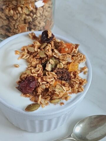 white bowl of yogurt topped with oil free granola placed on marble with a spoon next to it and a jar of granola behind it.