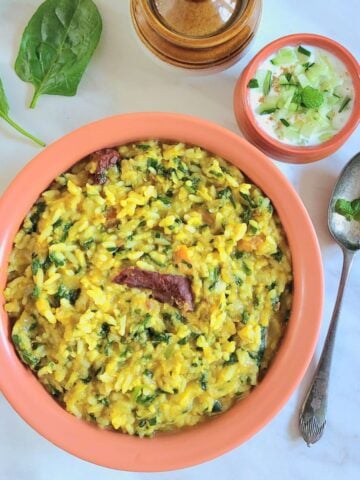 brown bowl filled with moong dal palak khichdi placed on a marble along with a spoon, cup of lassi and a pickle bowl.