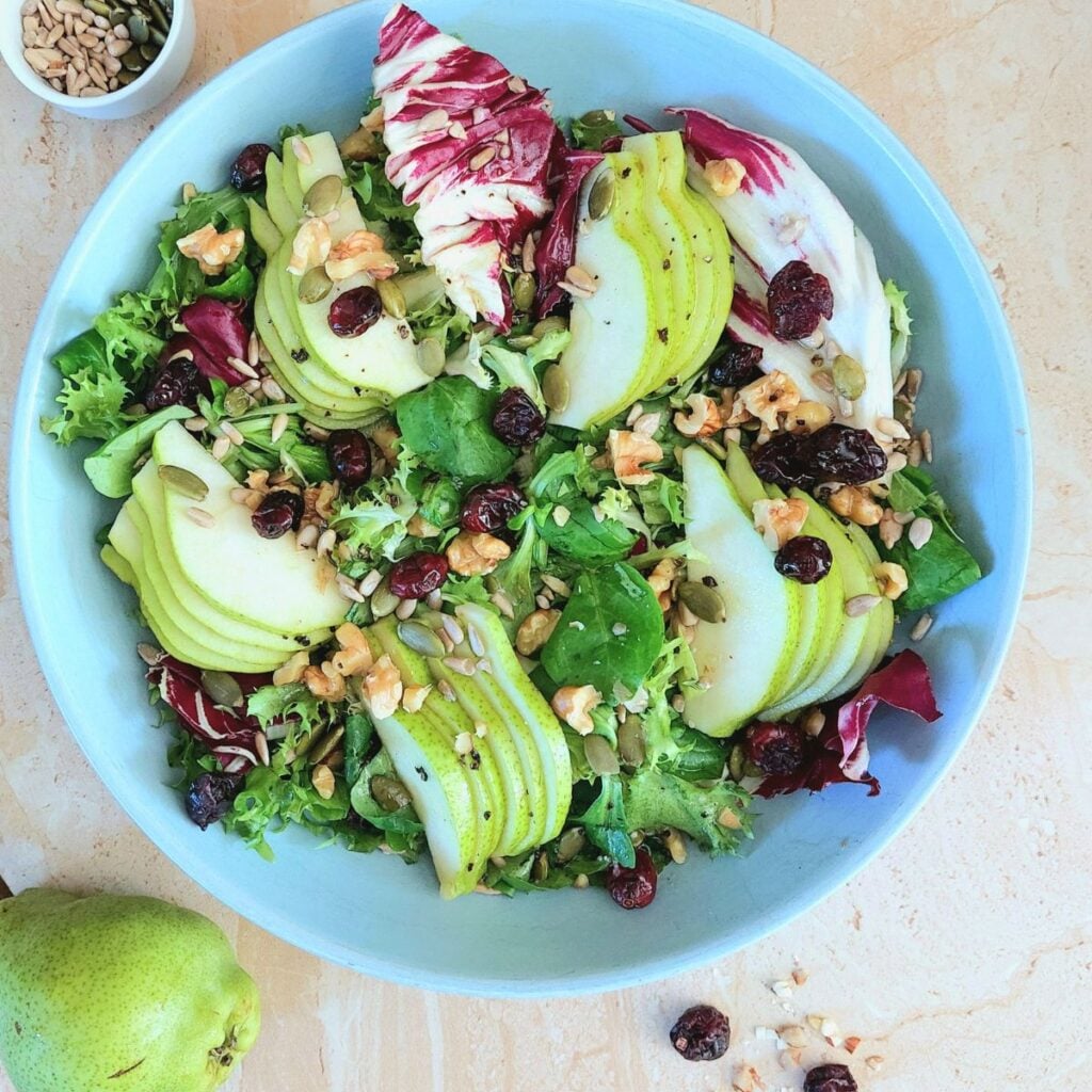 close up image of pear walnut cranberry salad in a blue bowl placed on a granite with a pear in the front and a bowl of seeds behind it.