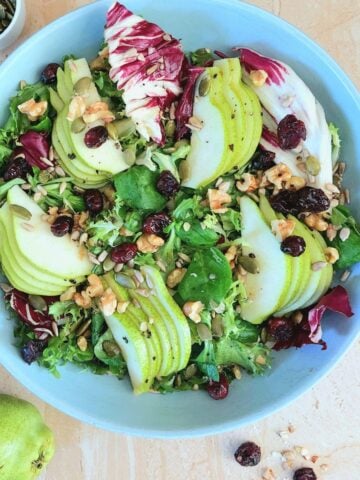 close up image of pear walnut cranberry salad in a blue bowl placed on a granite with a pear in the front and a bowl of seeds behind it.