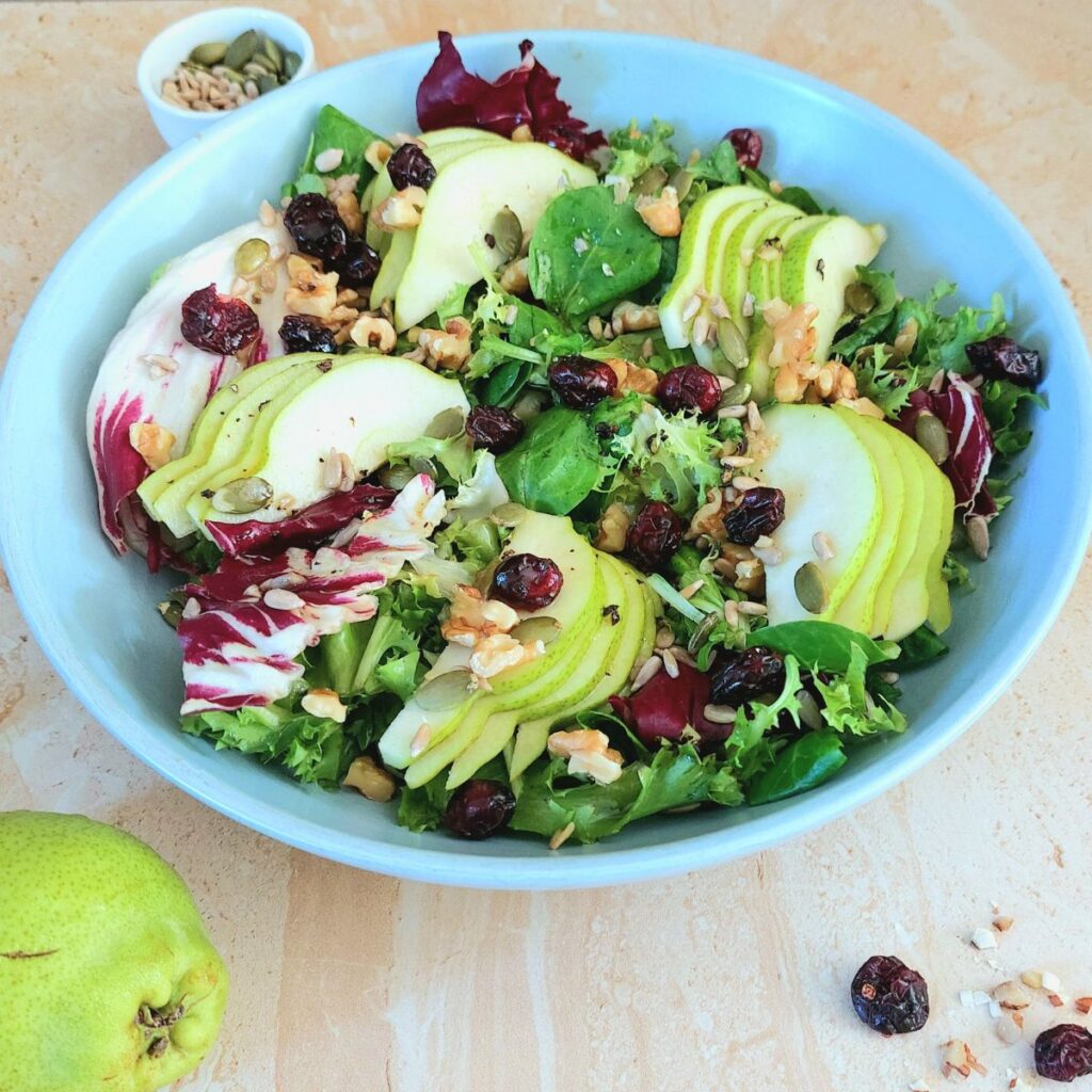 front view of the bowl of pear walnut green salad with a pear in the front and a bowl of seeds behind it.