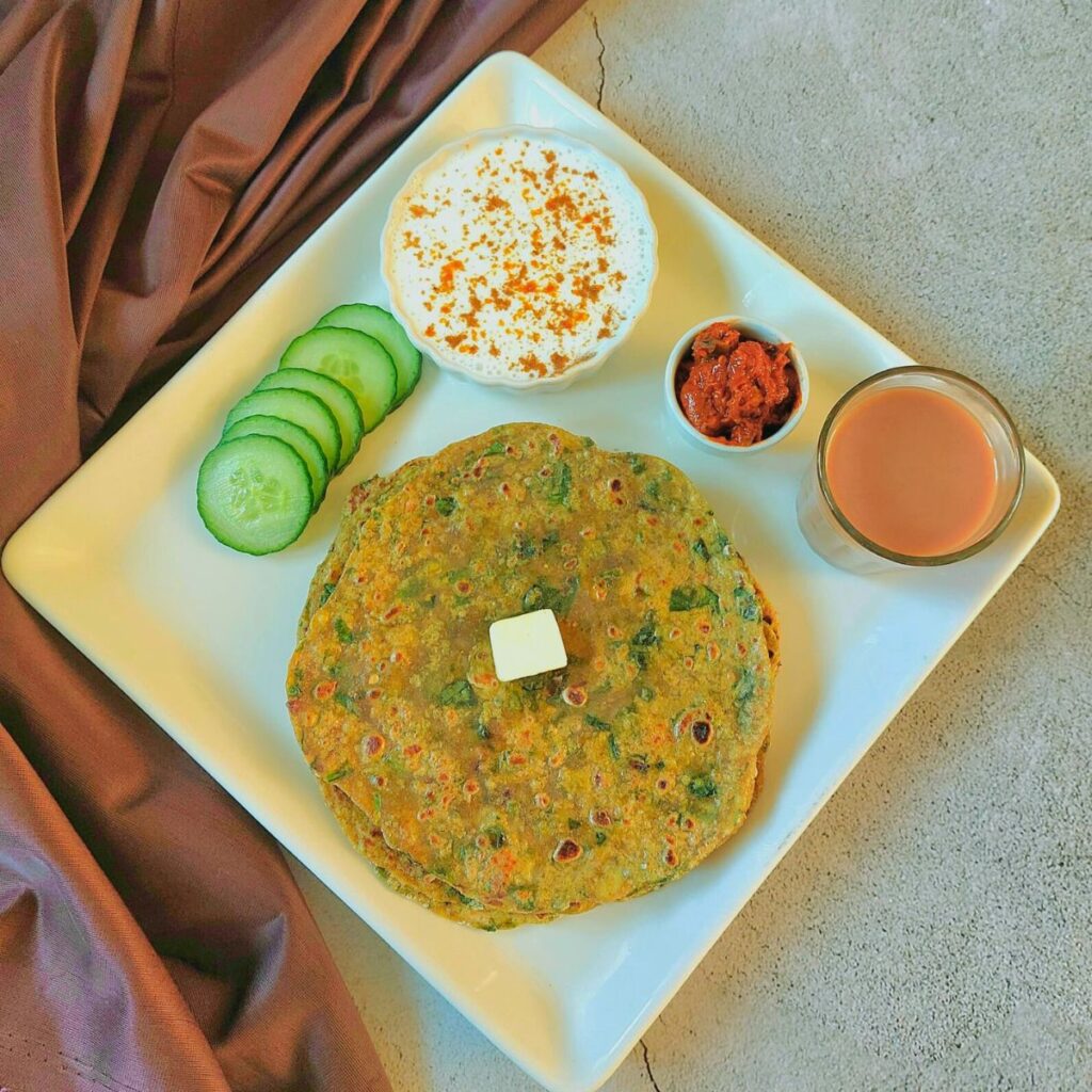 white square plate filled with stacked aloo palak roti with a dollop of ghee, glass of tea, cucumber slices, bowl of yogurt with spices garnished on top and a small bowl of pickle.