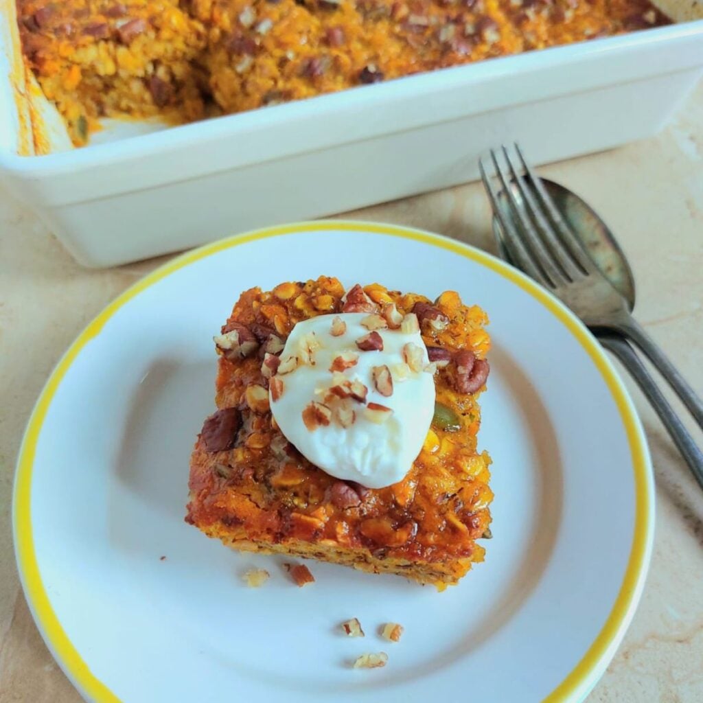 top view of slice of pumpkin baked oatmeal with yogurt nut topping on white plate with cutlery next to them.