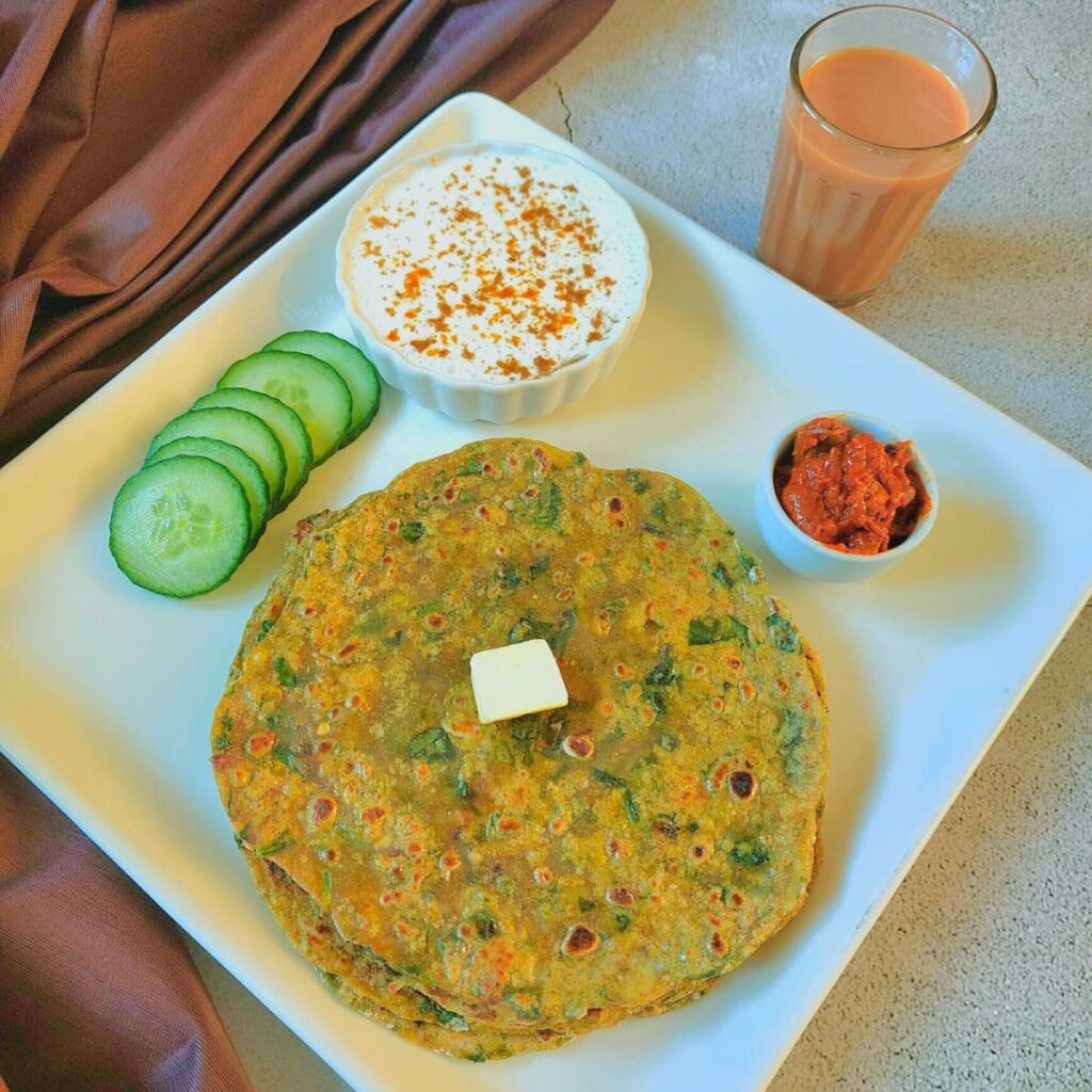 stack of aloo palak parathas placed on white plate along with cucumber slices, bowl of raita, pickle, glass of tea and a brown cloth next to plate.