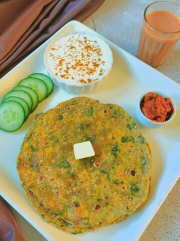stack of aloo palak parathas placed on white plate along with cucumber slices, bowl of raita, pickle, glass of tea and a brown cloth next to plate.