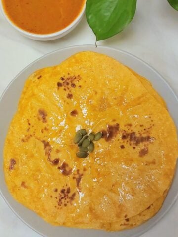 stack of pumpkin parathas on grey plate placed on marble along with a bowl of gravy