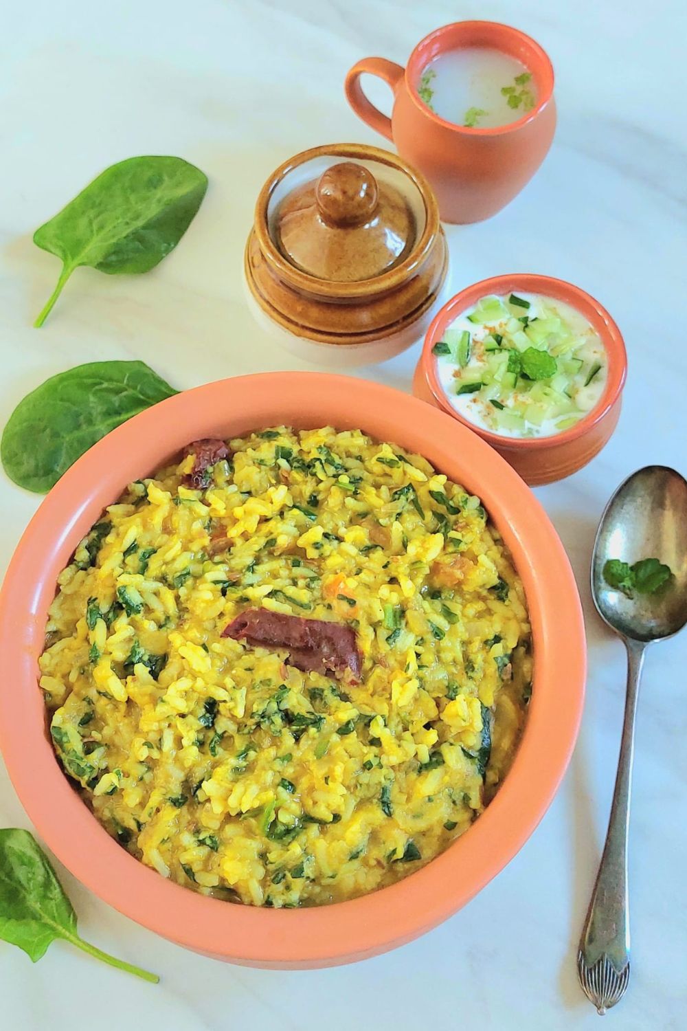 brown bowl of spinach lentil khichdi placed on a marble along with a spoon, glass of lassi, raita and a pickle container.