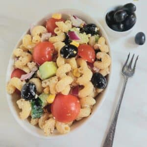 Greek vegetable pasta salad in a white bowl placed on marble along with a fork and a bowl of olives.