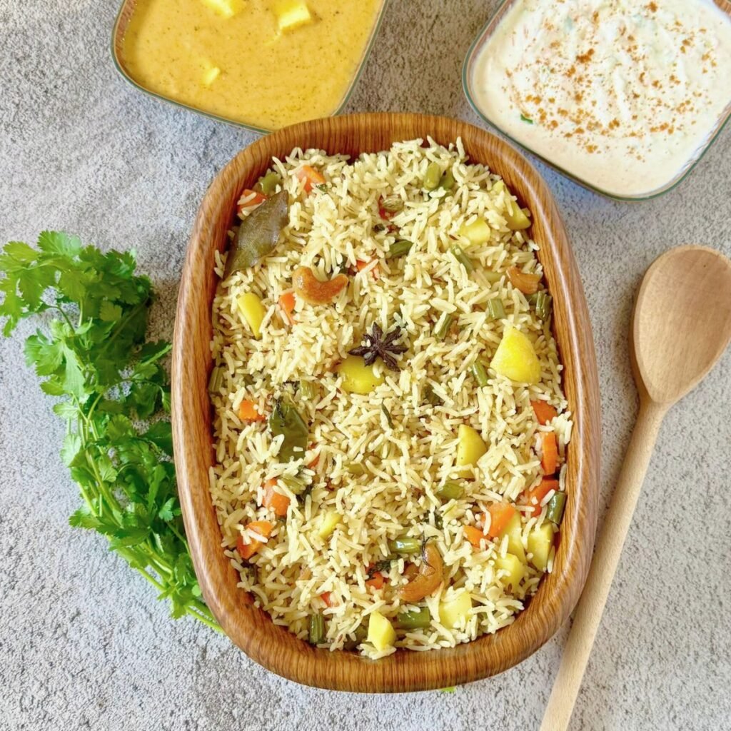 brown bowl of coconut milk pulao placed on black granite along with a bunch of coriander, wooden spoon, square bowls of yogurt and paneer curry