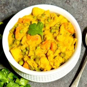top view of mixed vegetable kurma in a bowl placed on black tile along with a bunch of coriander leaves and a spoon.