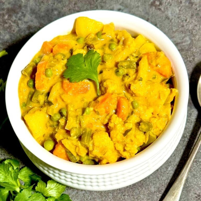 top view of mixed vegetable kurma in a bowl placed on black tile along with a bunch of coriander leaves and a spoon.