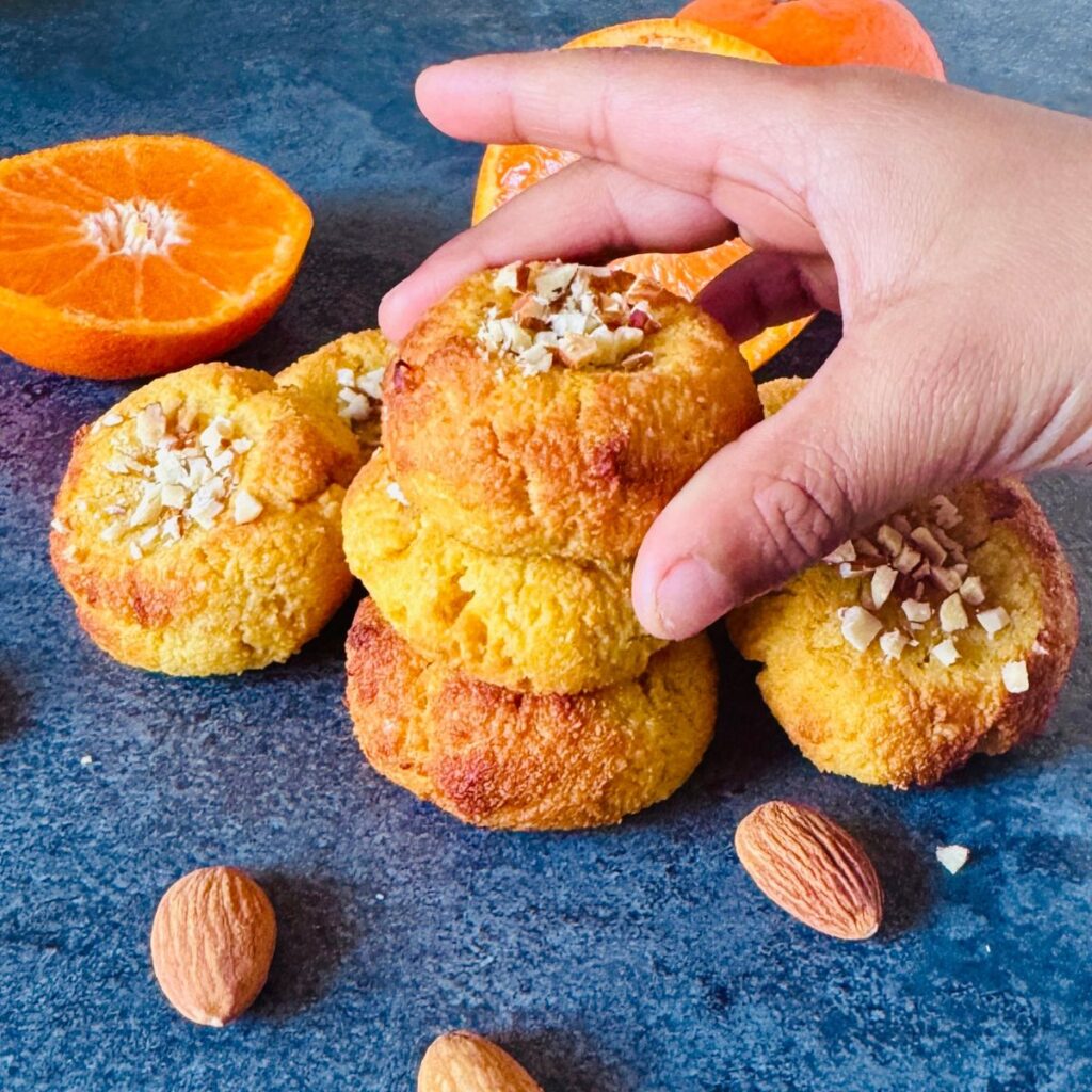 child picking orange almond cookie from a stack placed on a granite along with cut oranges.