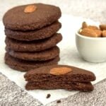 stack of 3 ingredient chocolate almond cookies placed on parchment paper along with with a half broken cookie and a pinch bowl of almonds.
