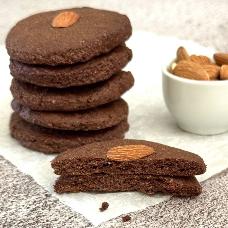 stack of 3 ingredient chocolate almond cookies placed on parchment paper along with with a half broken cookie and a pinch bowl of almonds.