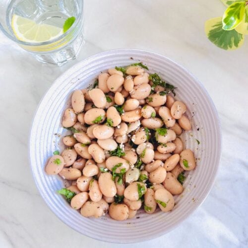 top view of a bowl of butter bean salad with garlic herb dressing placed on white marble along with a glass of water with lemon slice.