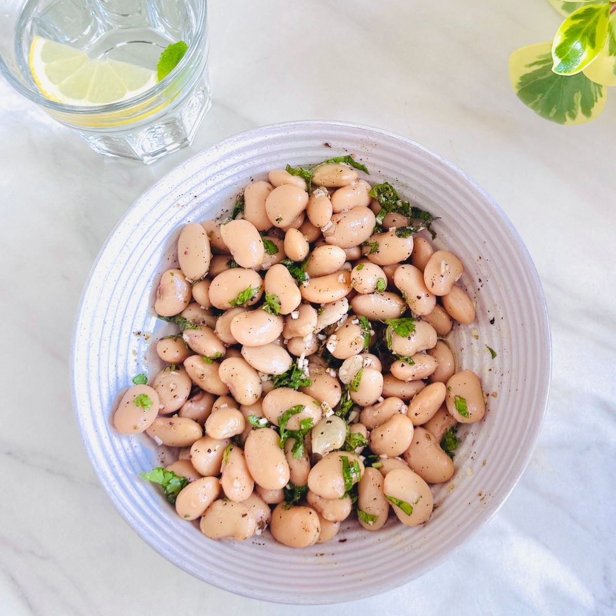 top view of a bowl of butter bean salad with garlic herb dressing placed on white marble along with a glass of water with lemon slice. 