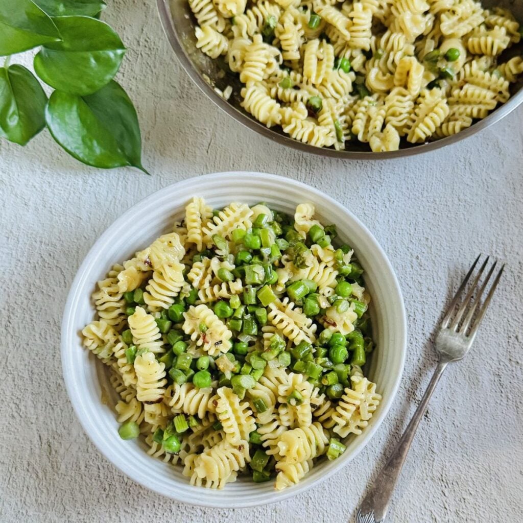 top view of a bowl of asparagus peas pasta, a fork and a steel pan of pasta.