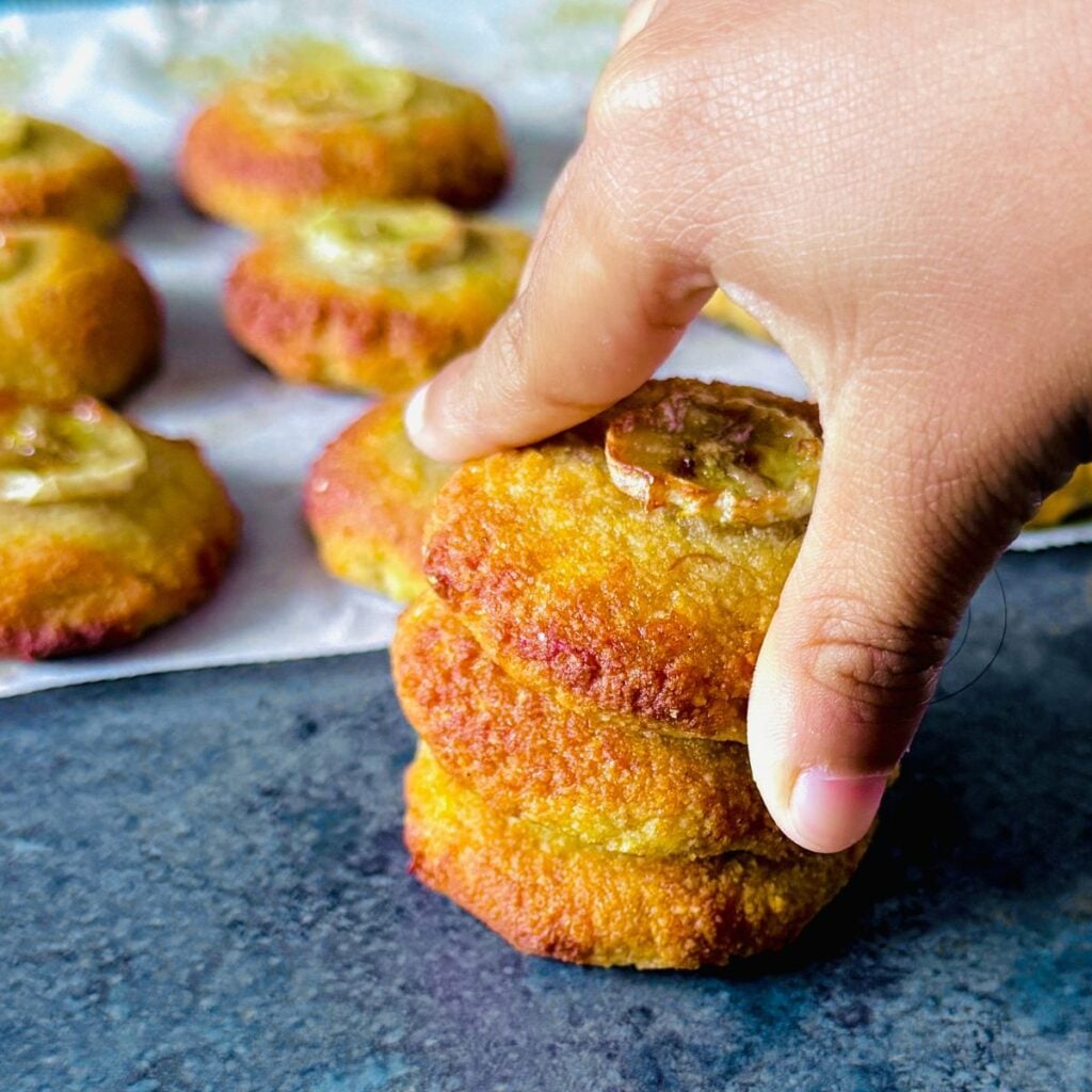 child holding banana almond cookie from the stack of cookies placed on granite.