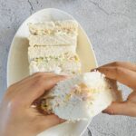 child holding sandwich with two hands on top of white plate of sandwiches
