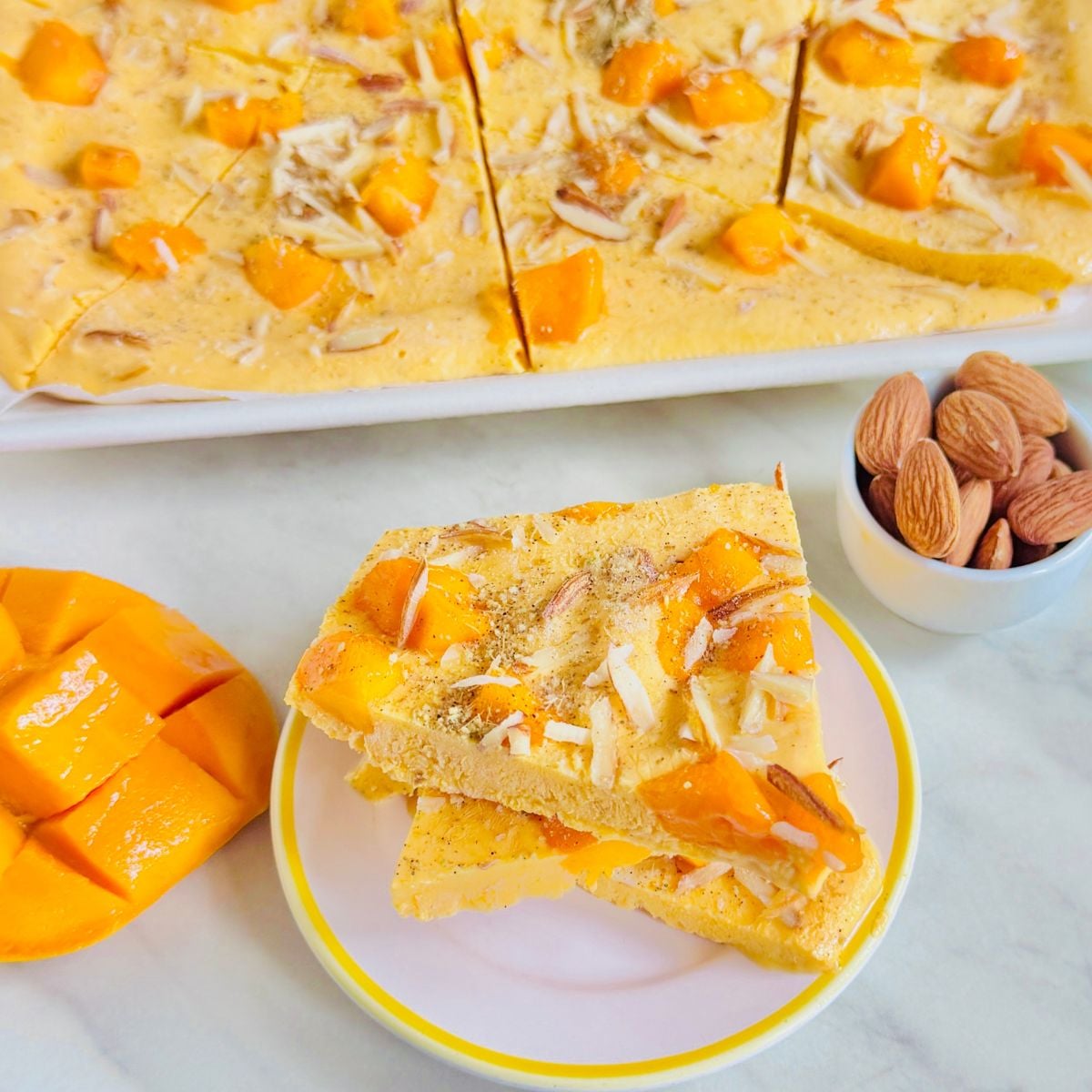 cardamom mango frozen yogurt bark placed on marble along with a tray of bark pieces, half cut mango and almonds.