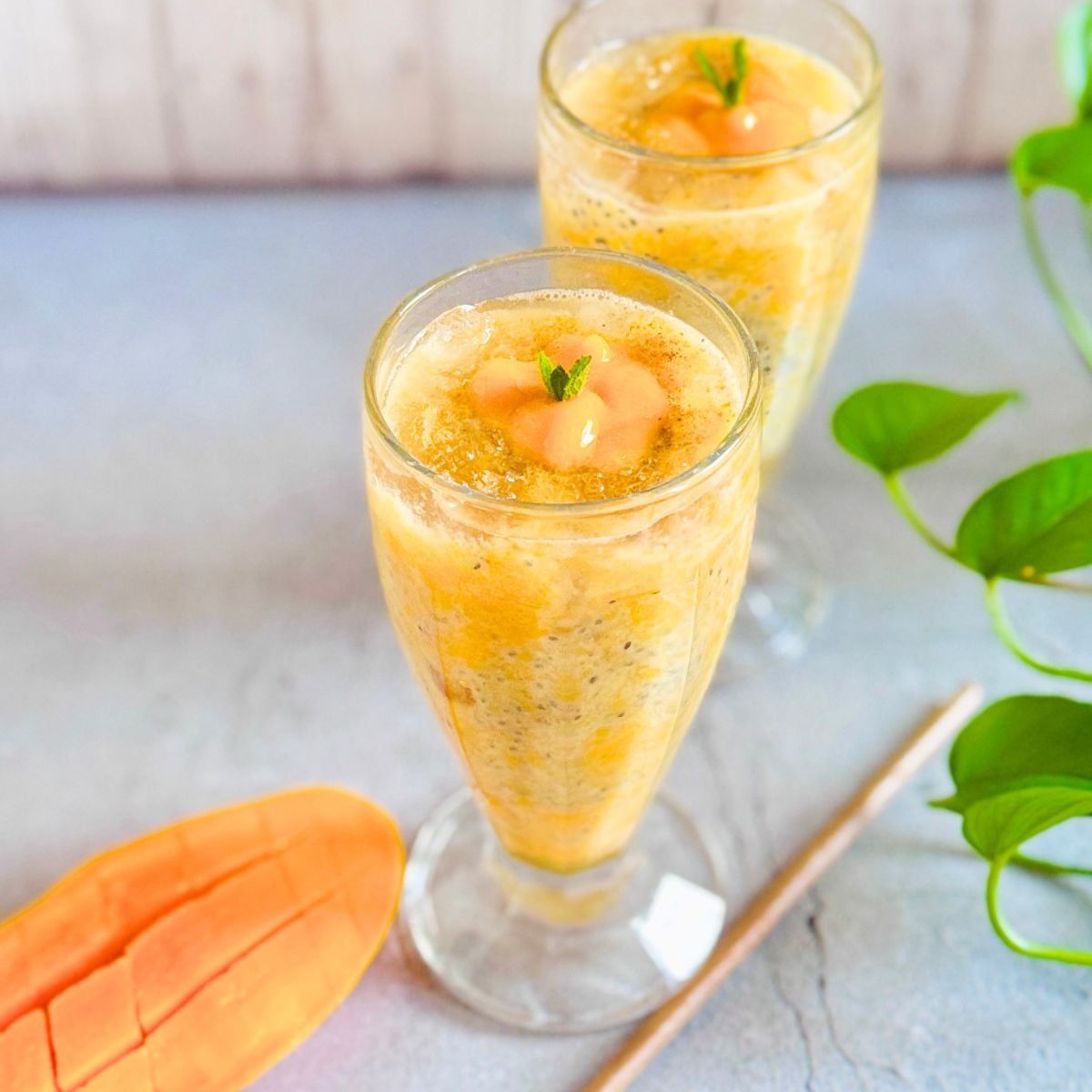 two glasses of mango gond katira sabja drink placed on a table along with an eco friendly coconut leaf straw and half cut mango.